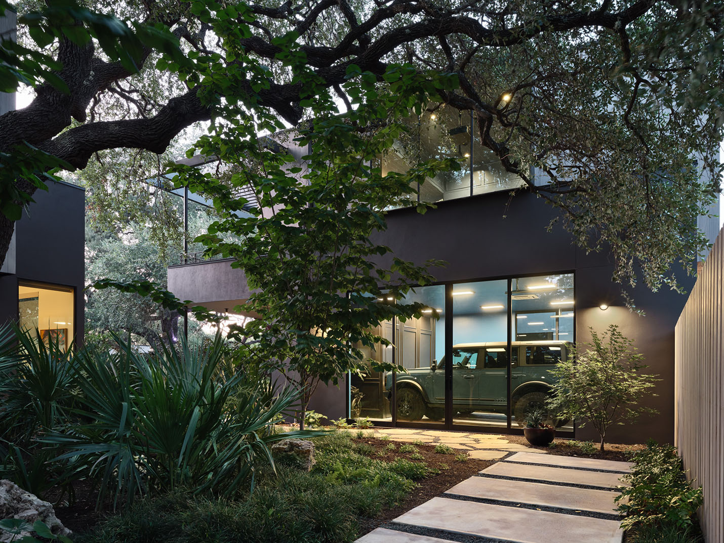 Evening view of a modern garage with glass doors and integrated lighting at the 5th Street House Addition in Austin, TX, surrounded by native trees and greenery.