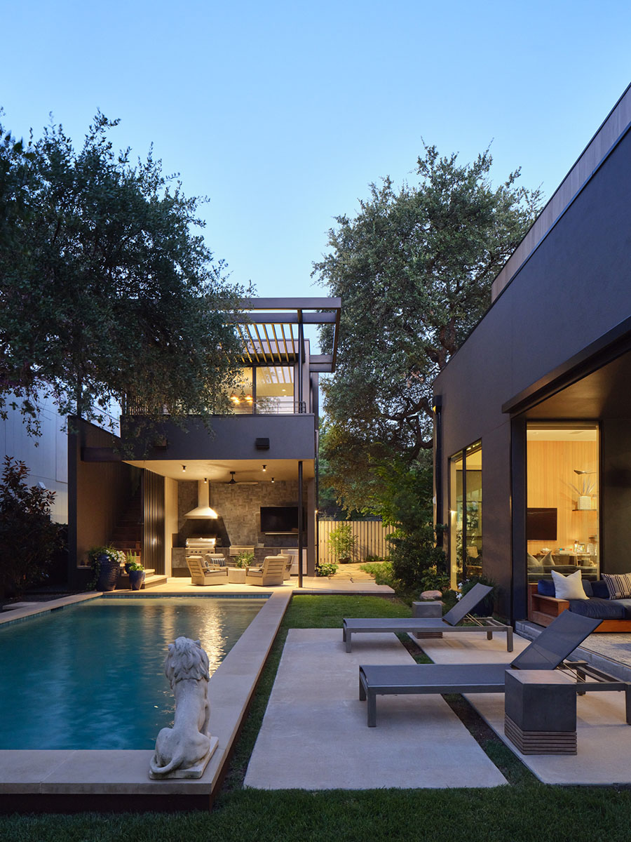 Dusk view of modern pool and outdoor living space with lounge seating, grill, and illuminated interiors at the 5th Street House Addition in Austin, TX.