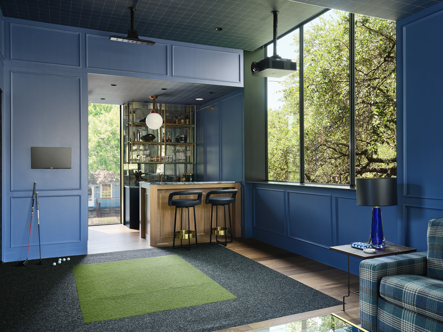 Stylish blue-paneled entertainment room with home bar, golf simulator mat, and large windows at the 5th Street House Addition in Austin, TX, built by Texas Construction Company.